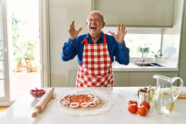 Senior man with grey hair cooking pizza at home kitchen crazy and mad shouting and yelling with aggressive expression and arms raised. frustration concept.