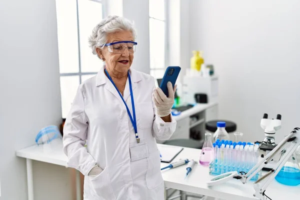 研究室でスマホを使って科学者の制服を着たシニア白髪の女性 — ストック写真
