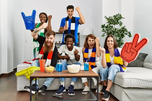 Group of friends supporting football team watching tv al home sitting on the sofa annoyed and frustrated shouting with anger, yelling crazy with anger and hand raised