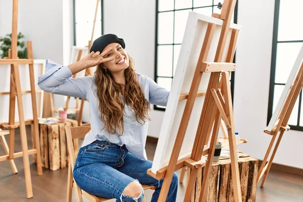 若いヒスパニックアーティストの女性は 顔の上に指で平和のシンボルを行うアートスタジオでキャンバス上で絵を描く 明るい勝利を示す笑顔 — ストック写真