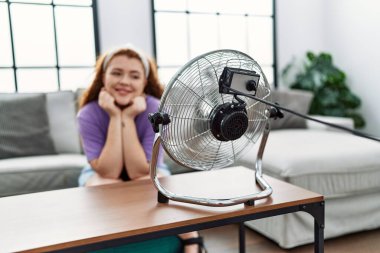 Genç kızıl saçlı kadın evinde elektrikli vantilatör kullandığından emin bir şekilde gülümsüyor.