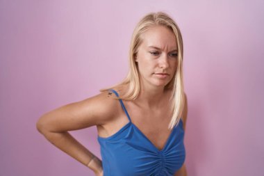 Pembe arka planda duran beyaz bir kadın sırt ağrısı çekiyor, elleriyle dokunuyor, kas ağrısı çekiyor. 