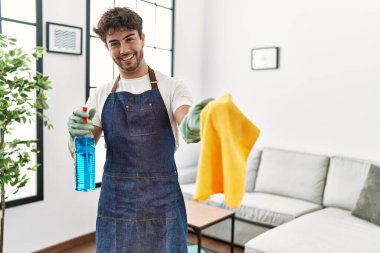Young hispanic man doing chores holding cleaning sprayer and cloth at home