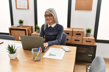 Ofiste çalışırken dizüstü bilgisayar kullanan, orta yaşlı, kır saçlı iş kadını.
