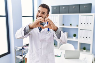 Doktor üniforması giyen yakışıklı İspanyol bir adam ve klinikte steteskop takıyor. Aşkla gülümsüyor. Elleriyle kalp sembolü ve şekli gösteriyor. romantik kavram. 
