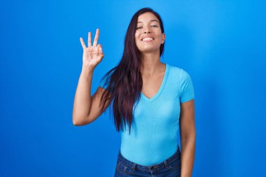 Mavi arka planda duran genç İspanyol kadın gülümsüyor ve el ve parmaklarıyla 
