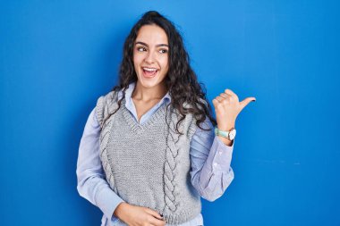 Mavi arka planda dikilen esmer kadın gülümsüyor, gülümsüyor ve başparmağıyla yan tarafa bakıyor.. 