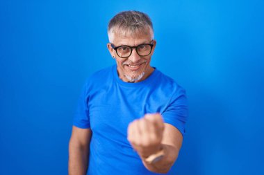 Mavi arka planda dikilen beyaz saçlı İspanyol bir adam, el kol hareketi yaparak, mutlu ve gülümseyen bir şekilde buraya gelir. 