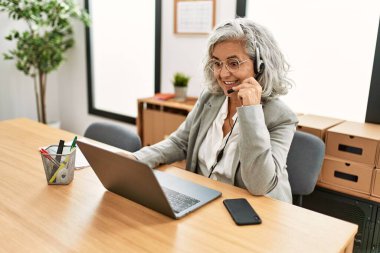Orta yaşlı, gri saçlı çağrı merkezi ajanı kadın ofiste çalışırken mutlu bir şekilde gülümsüyor..