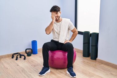 Sakallı İspanyol bir adam yoga odasında pilatese oturmuş bir gözünü eliyle kapatıyor, yüzünde kendine güvenen bir gülümseme ve sürpriz bir duygu var.. 