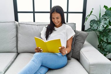 Genç Latin kadın gülümsüyor evde kendine güvenen kitap okuyor.