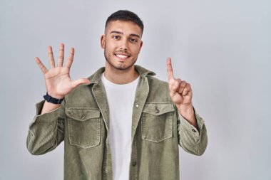 Genç İspanyol adam izole edilmiş arka planda duruyor. Altıncı parmaklarını gösterip işaret ederken kendinden emin ve mutlu bir şekilde gülümsüyor.. 