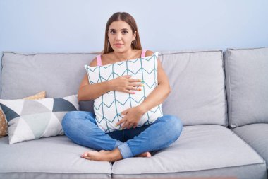 Young blonde woman hugging pillow sitting on the sofa clueless and confused expression. doubt concept. 