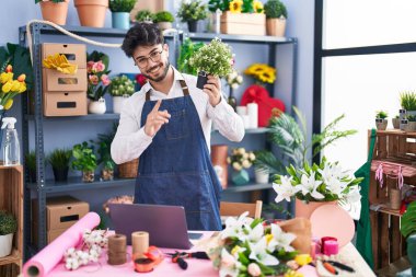 Young hispanic man florist using laptop holding plant at florist shop
