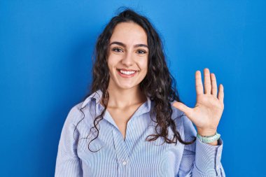 Mavi arka planda duran genç esmer kadın beş numarayı gösterip işaret ederken kendinden emin ve mutlu bir şekilde gülümsüyor.. 