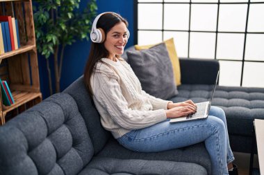Evdeki kanepede oturan dizüstü bilgisayar ve kulaklık kullanan genç bir kadın.