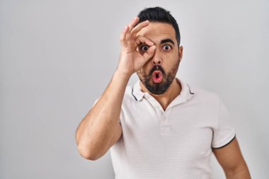 Sakallı, beyaz arka planda günlük giysiler giyen genç İspanyol adam şaşırmış yüz ifadesiyle şaşırmış, parmaklarından bakan bir jest yapıyor. İnançsız ifade. 