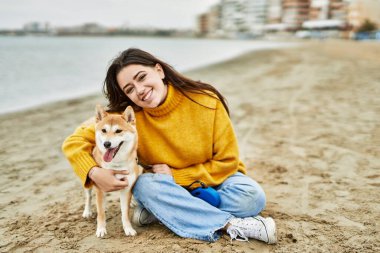 Güzel genç bir kadın sahilde mutlu Shiba Inu köpeğine sarılıyor.