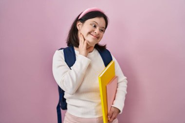 Down sendromlu bir kadın sırt çantası takıyor ve elinde kitaplar tutuyor. Gülümsüyor, saçlarına dokunuyor, el hareketi yapıyor, çekici ve modaya uygun poz veriyor. 