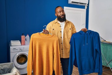 Çamaşır odasında askı üzerinde temiz giysiler tutan Afro-Amerikan bir adam gülümsüyor ve başka tarafa bakıyor.. 