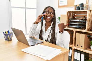 Ofiste örgü ören siyah bir kadın telefonda gülen bir yüzle gülümseyen ve baş parmağıyla yan tarafı gösteren.. 