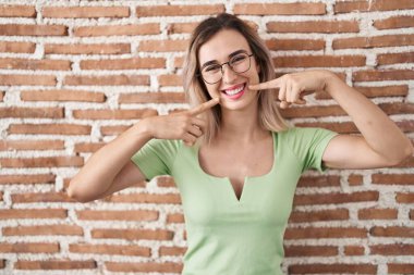 Tuğla duvarın üzerinde duran genç ve güzel bir kadın neşeli bir şekilde gülümsüyor ve parmakları ve ağızlarıyla işaret ediyor. diş sağlığı kavramı. 