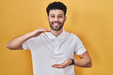 Arap adam sarı arka planda duruyor. Elleri büyük ve büyük bir işaret, ölçü sembolü gösteriyor. Kameraya bakıp gülümsüyor. ölçüm kavramı. 