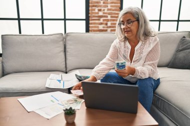 Orta yaşlı kadın kumar paraları sayıyor ve evde dizüstü bilgisayar kullanıyor.