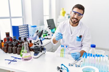 Laboratuvarda pipet ve test tüpü kullanarak bilim adamı üniforması giyen İspanyol genç bir adam.