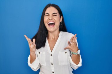 Mavi arka planda duran genç İspanyol kadın başarı için çıldırıyor ve deliriyor kollarını kaldırıyor ve gözleri kapalı heyecanla bağırıyor. Kazanan konsept 