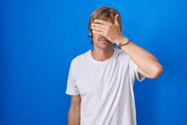 Orta yaşlı bir adam mavi arka planda duruyor. Gözleri elle kaplıyor, ciddi ve üzgün görünüyor. Görme, saklama ve reddetme konsepti 