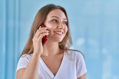 Sarışın, kendine güvenen genç bir kadın izole edilmiş mavi duvar arka planında akıllı telefondan konuşuyor.