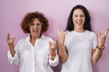 İspanyol anne ve kızı pembe arka plan üzerine günlük beyaz tişört giyip çılgın bir ifadeyle elleri havada rock sembolü yaparak bağırıyorlar. Müzik yıldızı. ağır müzik konsepti. 