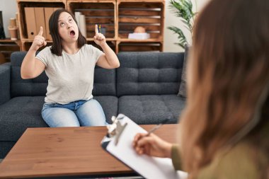 Down sendromlu İspanyol kız terapi yapıyor. Şaşırmış ve şaşırmış bir şekilde yukarı bakıyor ve parmaklarıyla işaret ediyor ve kollarını kaldırıyor.. 