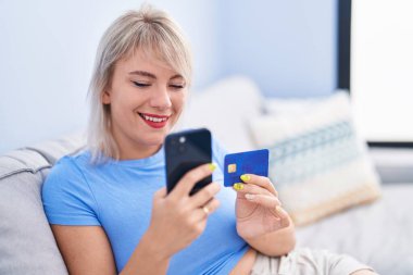 Akıllı telefon ve kredi kartı kullanan genç sarışın kadın evdeki koltukta oturuyor.