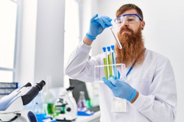 Laboratuvarda test tüpleri tutan bilim adamı üniforması giyen genç kızıl saçlı adam.