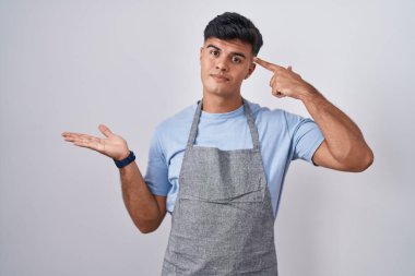 Beyaz arkaplan üzerine önlük giymiş İspanyol genç bir adam avuç içinin açık olmasından rahatsız ve kafası karışmış bir şekilde fotokopi alanını ve alnını işaret ediyor. Bunu bir düşün.. 
