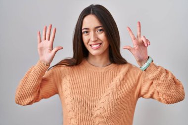 Genç esmer kadın beyaz arka planda duruyor. Sekiz numaralı parmaklarıyla işaret ediyor ve gülümsüyor. Kendinden emin ve mutlu.. 