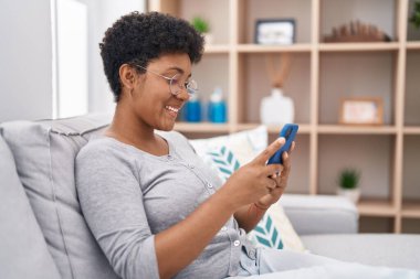 Evdeki koltukta oturan Afro-Amerikan kadın akıllı telefon kullanıyor