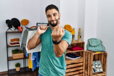 Young handsome man with beard holding shopping bags at retail shop showing middle finger, impolite and rude fuck off expression 