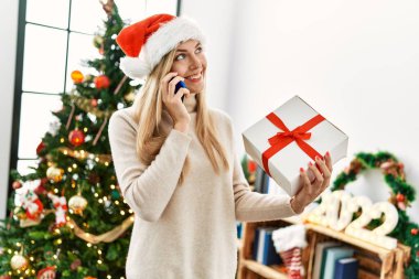 Genç sarışın kadın akıllı telefondan konuşuyor. Elinde Noel hediyesi var.