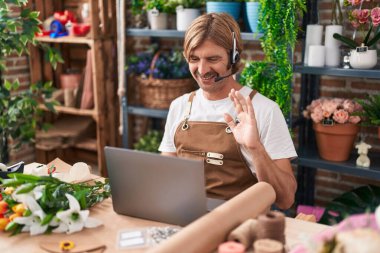Çiçekçide çalışan beyaz bıyıklı bir adam video çekiyor. Olumlu ve mutlu görünüyor. Kendine güvenen bir gülümsemeyle dişlerini gösteriyor. 