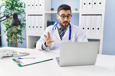 Doktor üniforması giyen genç İspanyol bir adam klinikten video çağrısı alıyor.