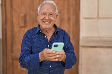 Yaşlı, gri saçlı, kendine güvenen, caddede akıllı telefon kullanan bir adam.