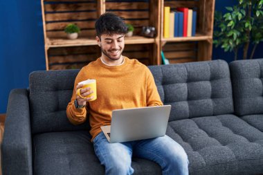 Young hispanic man using laptop and drinking coffee sitting on sofa at home
