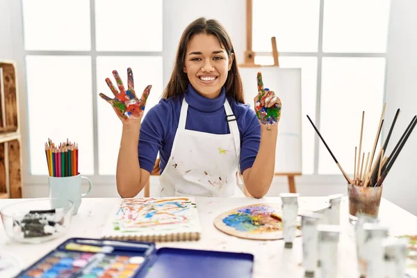 Young brunette woman at art studio with painted hands showing and pointing up with fingers number six while smiling confident and happy.