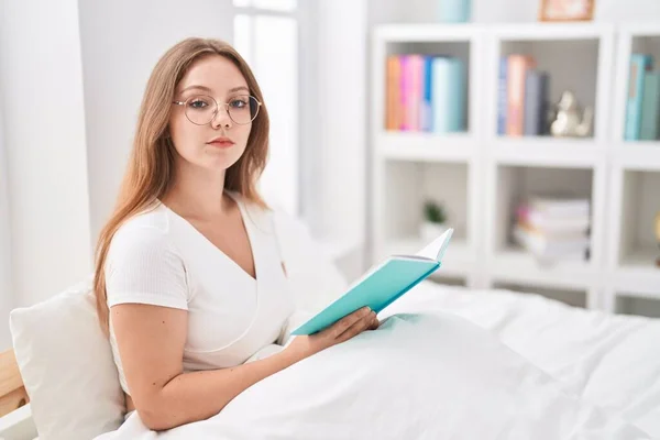 Young caucasian woman sitting on the bed at home reading a book thinking attitude and sober expression looking self confident