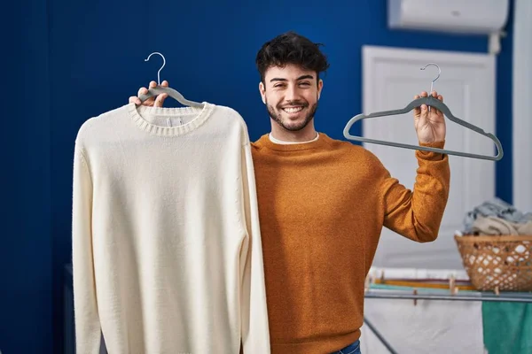 在洗衣房 一个留着胡子的西班牙男人拿着毛衣挂在衣架上笑着 脸上带着快乐而冷静的笑容 显示牙齿 — 图库照片
