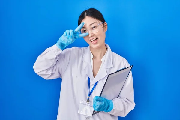 Chinese young woman working at scientist laboratory doing peace symbol with fingers over face, smiling cheerful showing victory