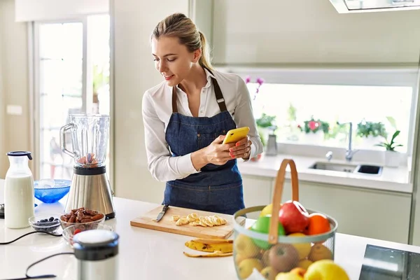 年轻的金发女人在厨房用智能手机切香蕉 — 图库照片